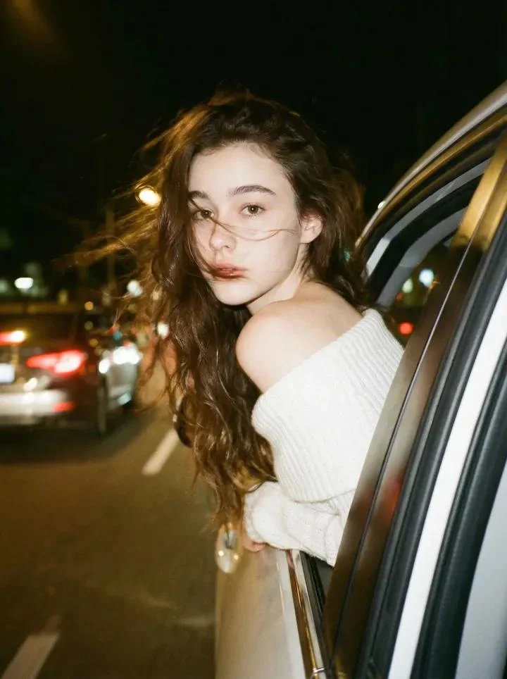 AI image: Create a photo of a person leaning their back out the window car like a dark dreamy blurry vintage windy night wearing a off shoulder white knitted top. With brown long wavy hair. Keep the facial details correctly.