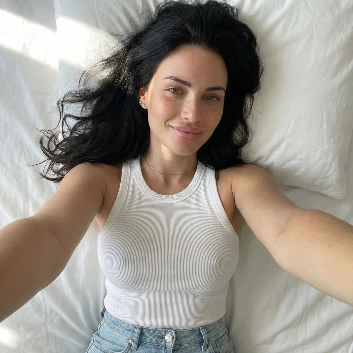 AI image: Young woman lying on back taking selfie with right arm extended upward. Soft smile, relaxed natural expression, looking toward camera. Smooth warm sunlit skin glow with visible freckles on nose and cheeks. Medium brown long hair, loose spread on white pillow around head. Light blue/green eyes with subtle eyeliner. White ribbed form-fitting scoop neck tank top. Natural sunlight from upper left creating warm highlights and soft shadows. Top-down selfie angle, close-up of face.